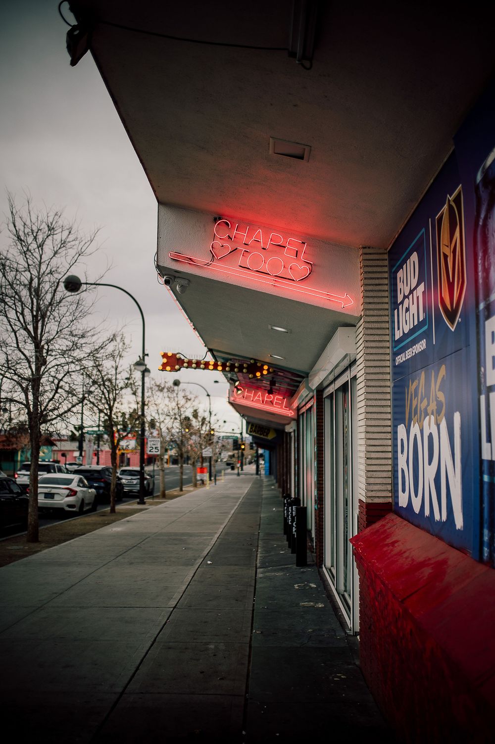 Urban storefront with red neon signage and stark shadows cast along empty sidewalk during dusk hours.