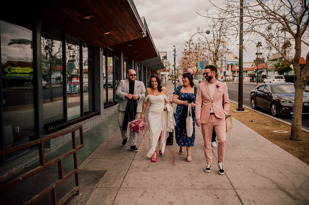 A group of well-dressed people walking together on an urban sidewalk outside storefronts on a sunny day.