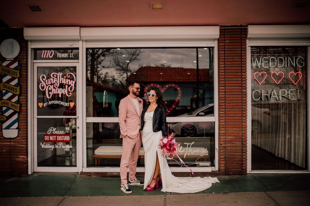 A couple poses in front of a brick storefront at night with illuminated window displays.