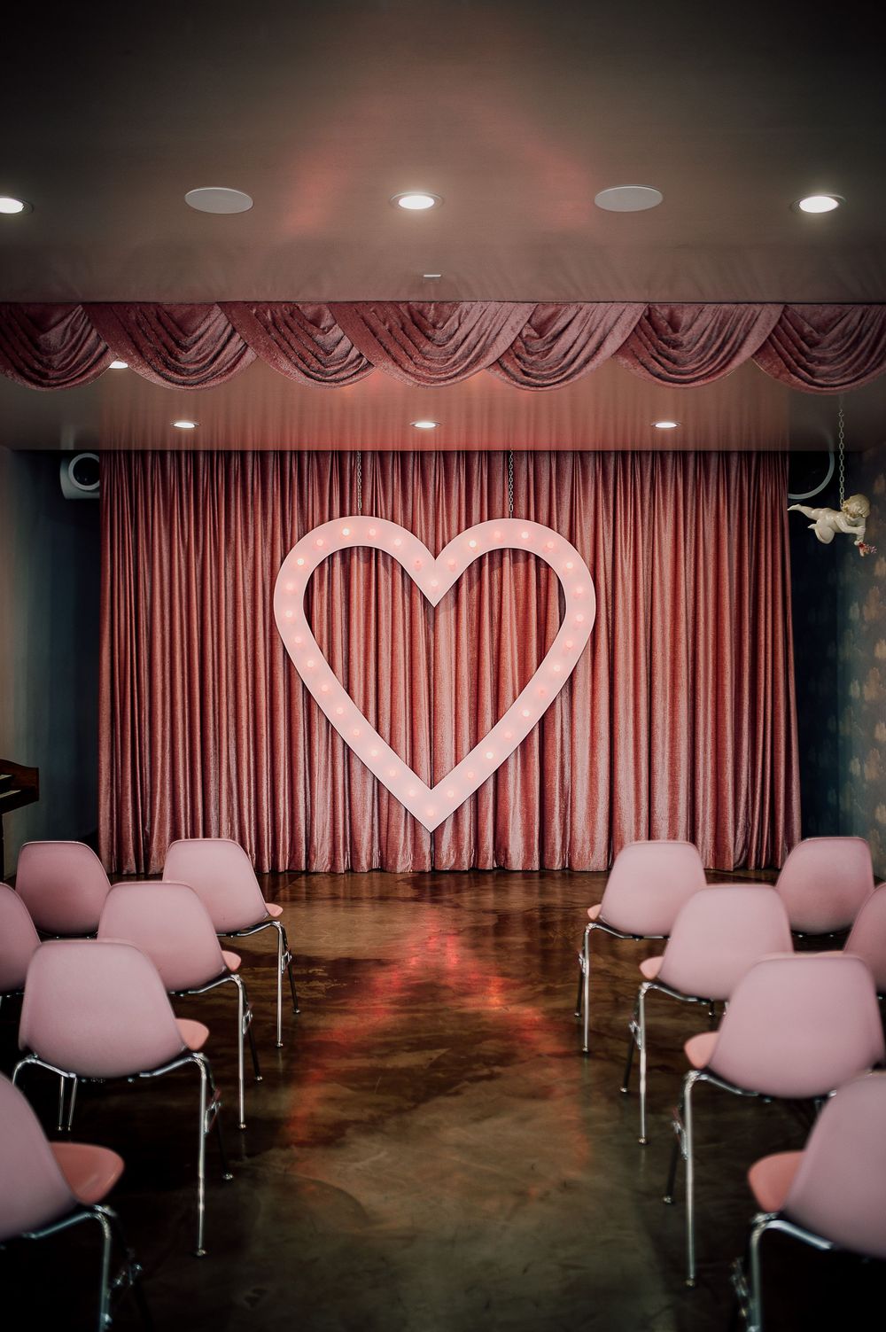 Pink chairs arranged in rows face a heart-shaped light display on a curtained stage in a dimly lit event space.