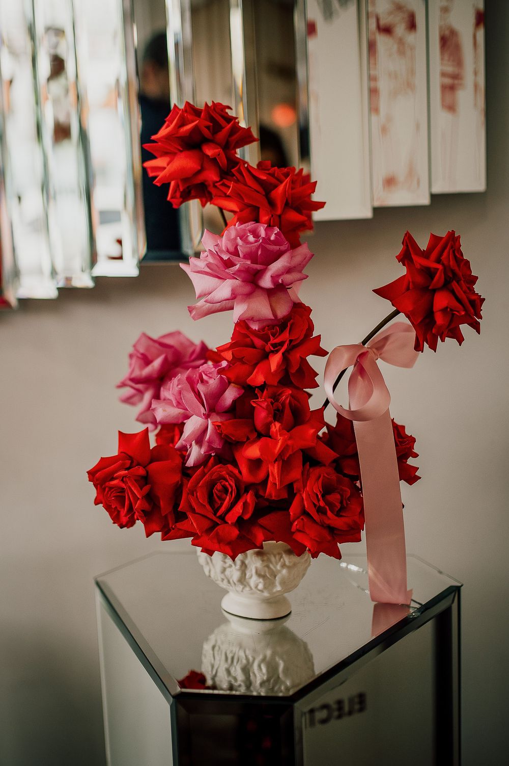 Red and pink carnations arranged in a white ceramic vase on a reflective surface with papers hanging in the background.