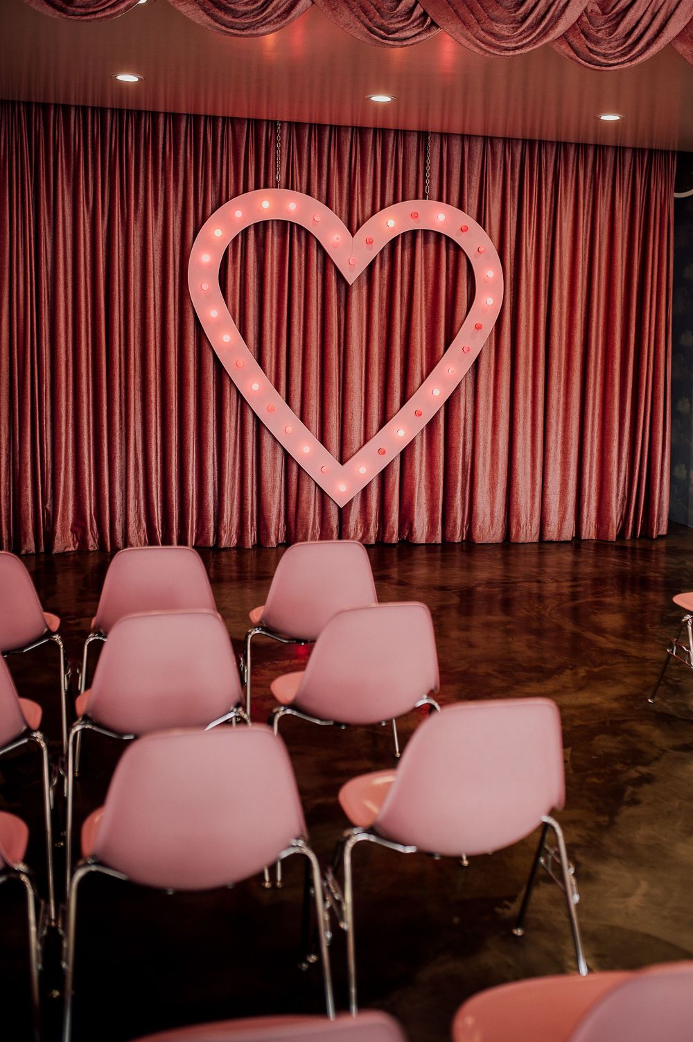 Pink heart decorations hang above modern white chairs in a dimly lit room with dark curtains and floors.