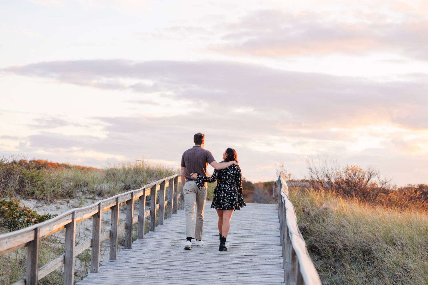 People walking on a wooden boardwalk path along a beach during sunset with coastal vegetation visible.