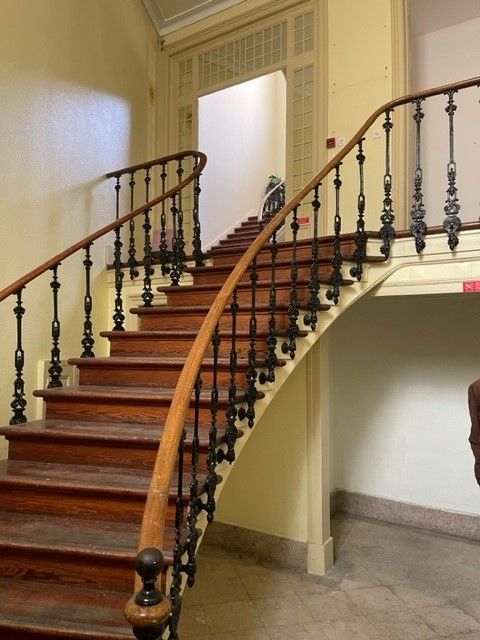 Elegant curved wooden staircase with ornate black iron balusters and polished handrail in a historic building interior.