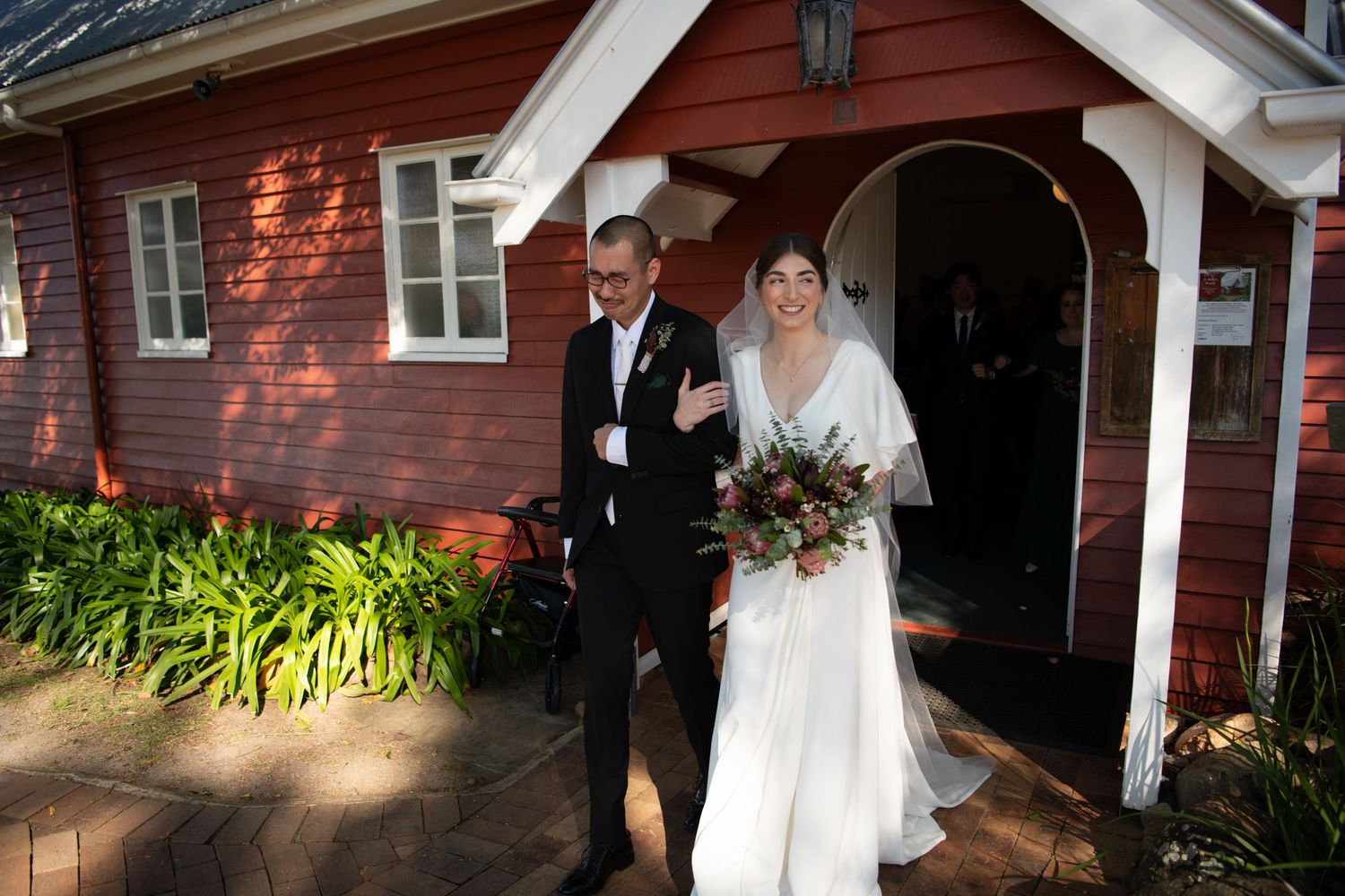 Bride in white gown walks with suited man from red wooden chapel after wedding ceremony.