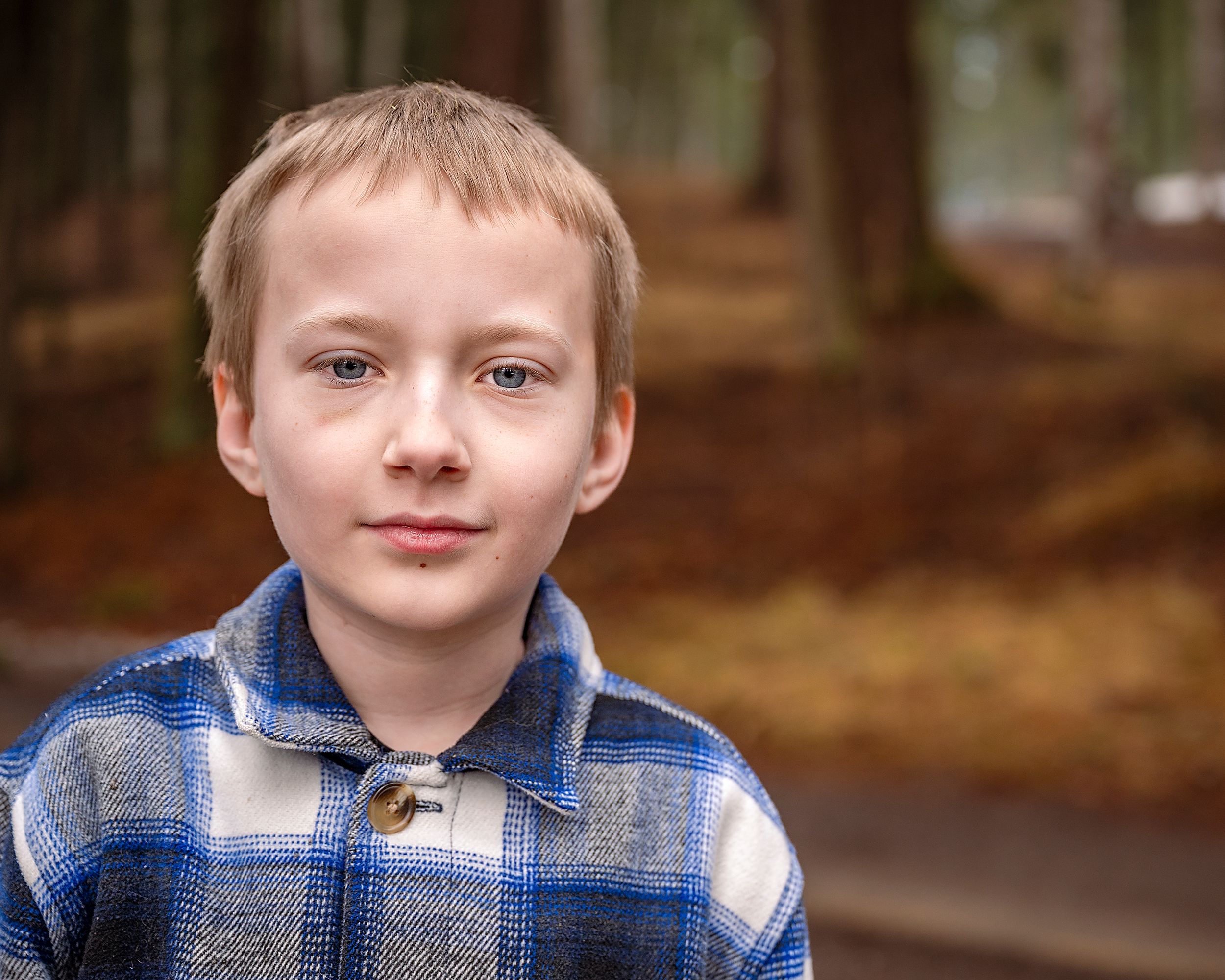 Young person wearing a blue and gray plaid flannel shirt poses outdoors in a wooded forest setting.