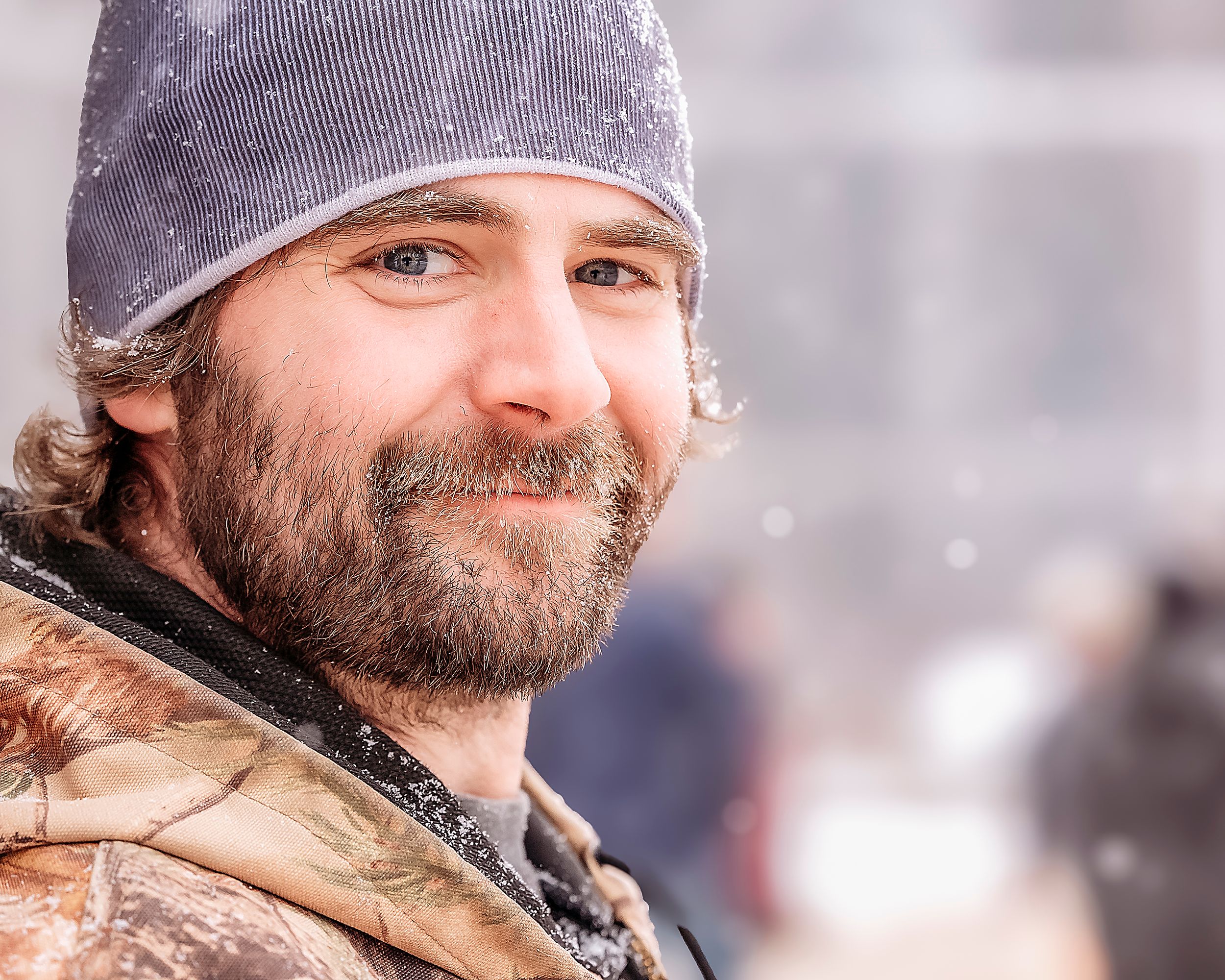 A bearded person wearing a winter hat smiles warmly at the camera while outdoors in snowy weather.