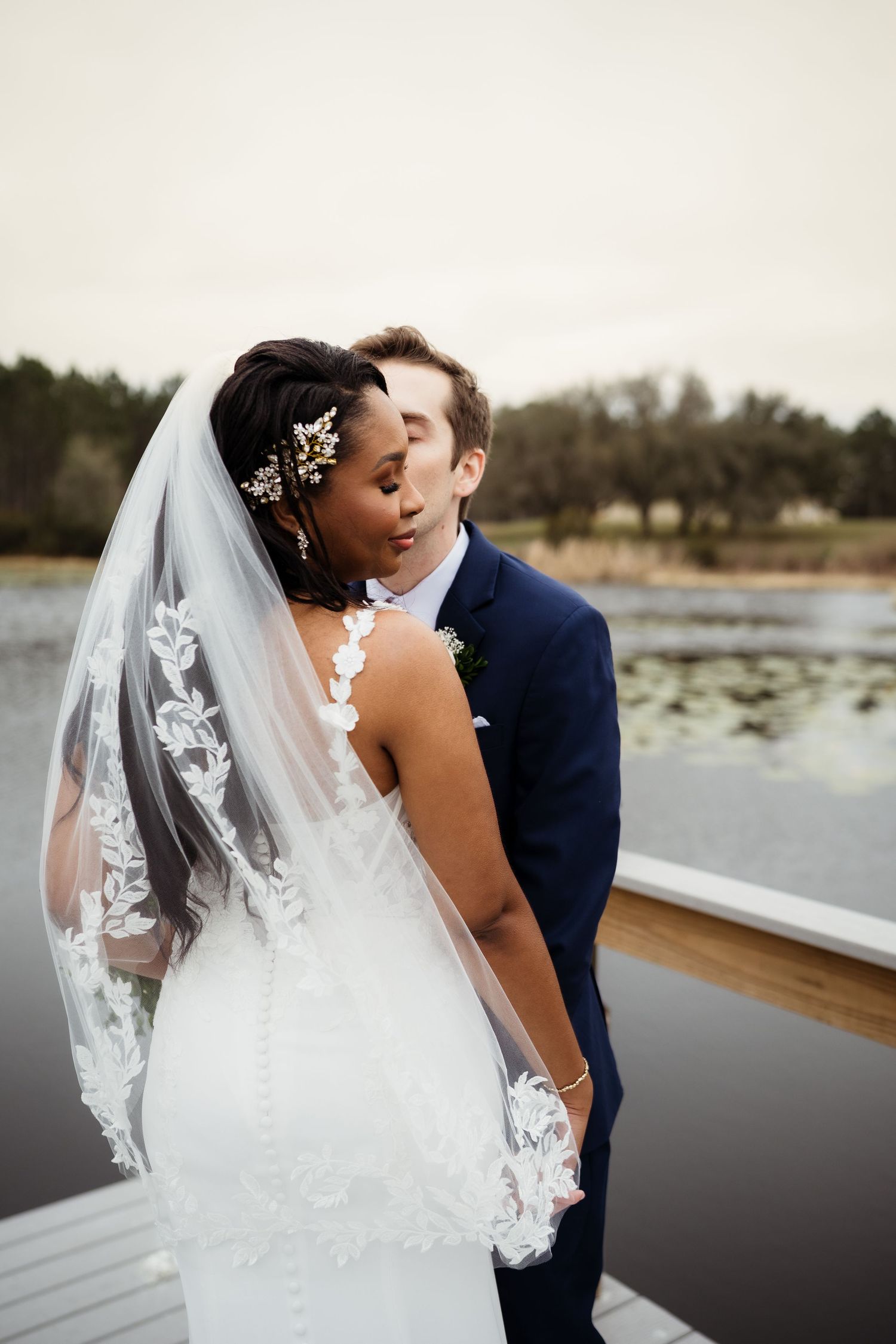 A romantic wedding couple embraces on a dock by a serene lake, with the bride wearing a lace veil and white dress.