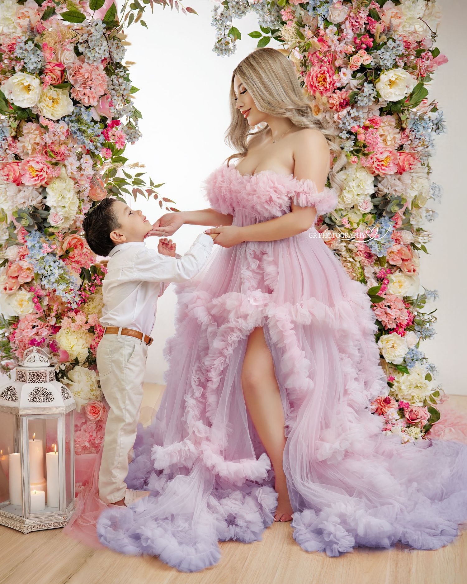 A dreamy maternity photo with a pink tulle gown against a floral arch backdrop with pink and white blooms and lantern decor.