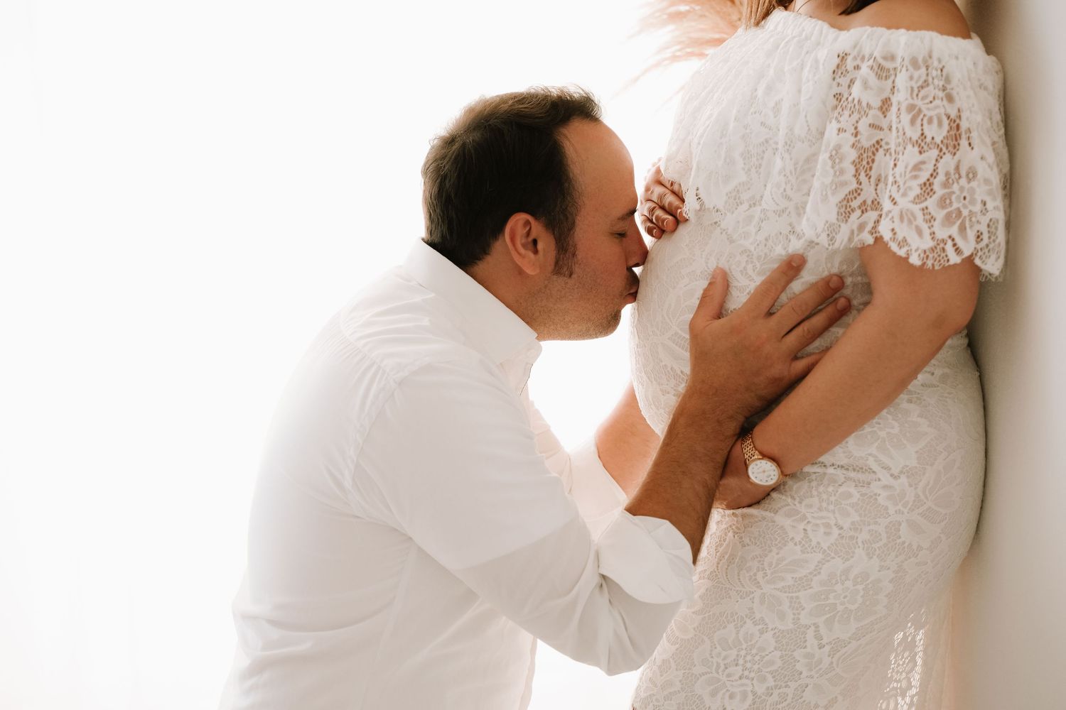 Man in white shirt kissing pregnant woman's belly in a bright, minimalist setting.