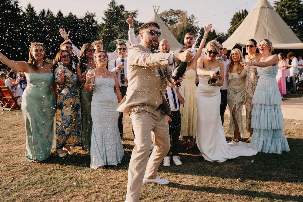 Wedding party celebrates joyfully outdoors with raised arms during sunset at a rustic outdoor reception.