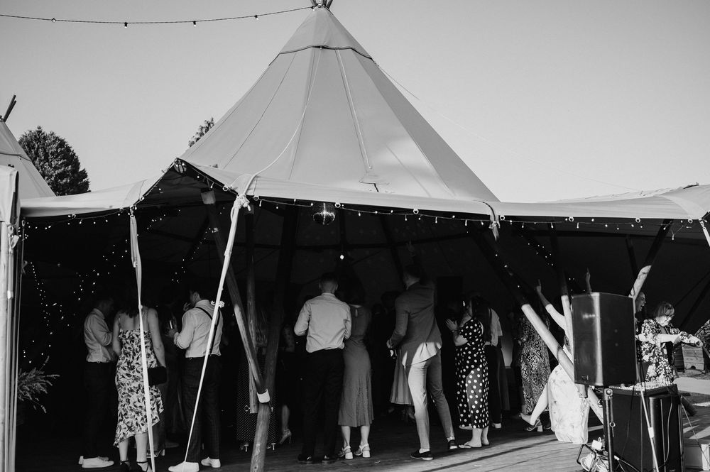 Black and white photo of a large wedding reception tent with string lights and guests mingling.