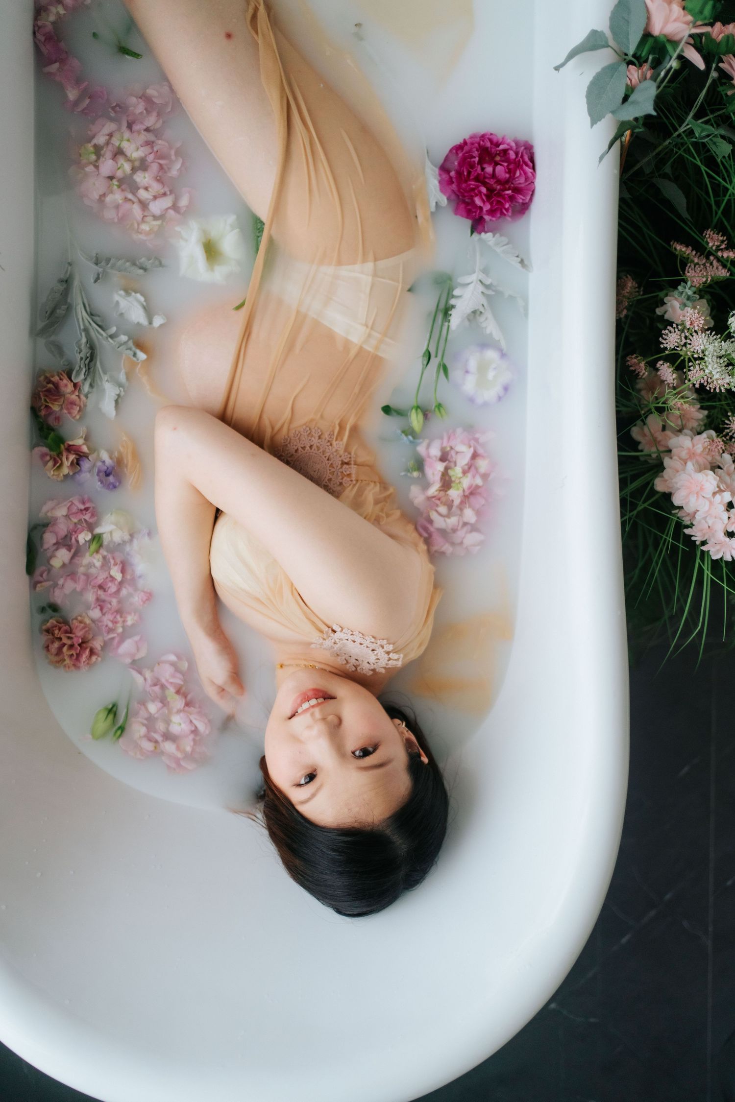Ethereal milk bath photography with delicate pink flowers floating around a figure in a bathtub.空靈的牛奶浴攝影作品，嬌嫩的粉紅色花朵圍繞著浴缸中的人物漂浮。