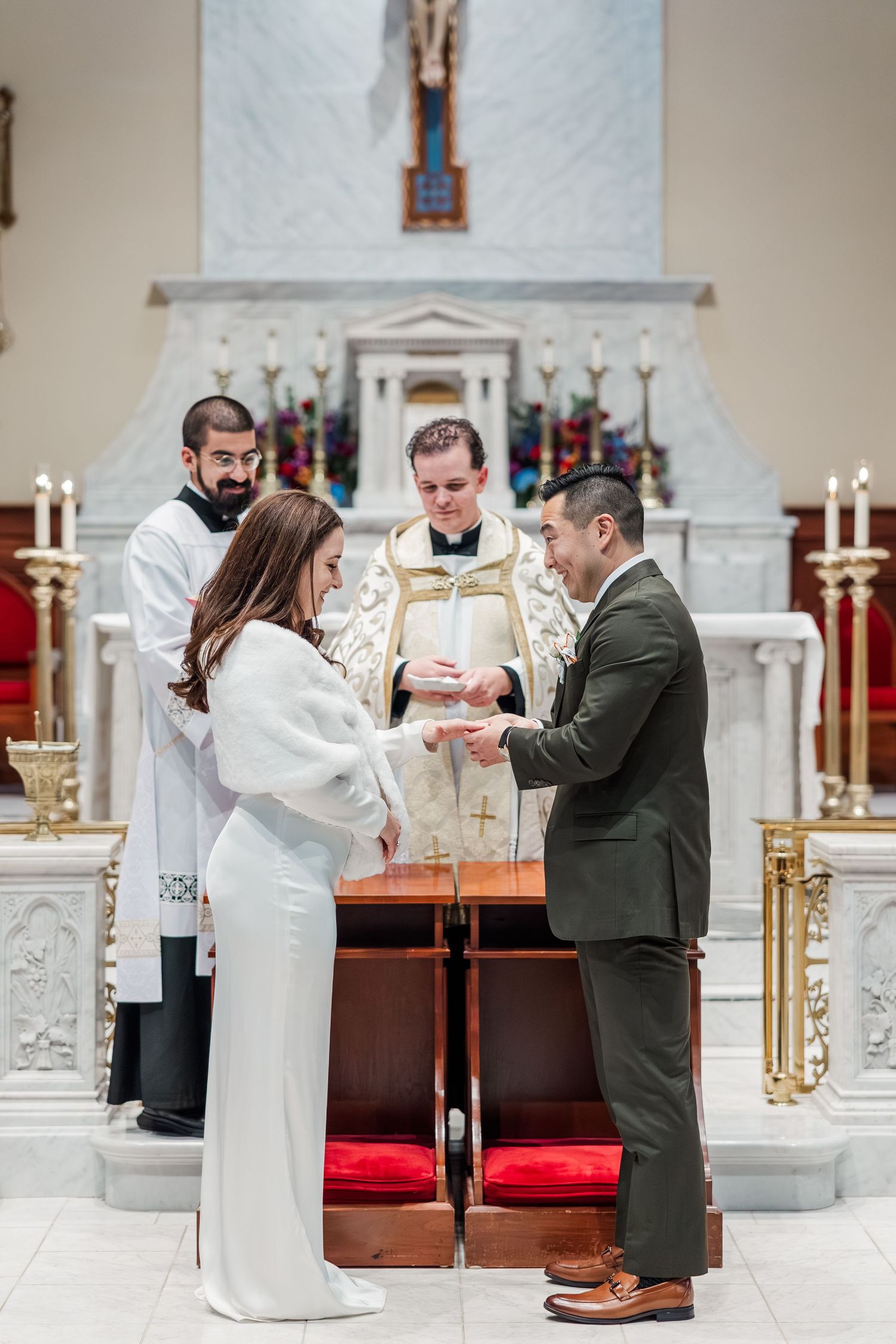 A couple exchanges rings during their wedding ceremony in a traditional white church with ornate decorations.