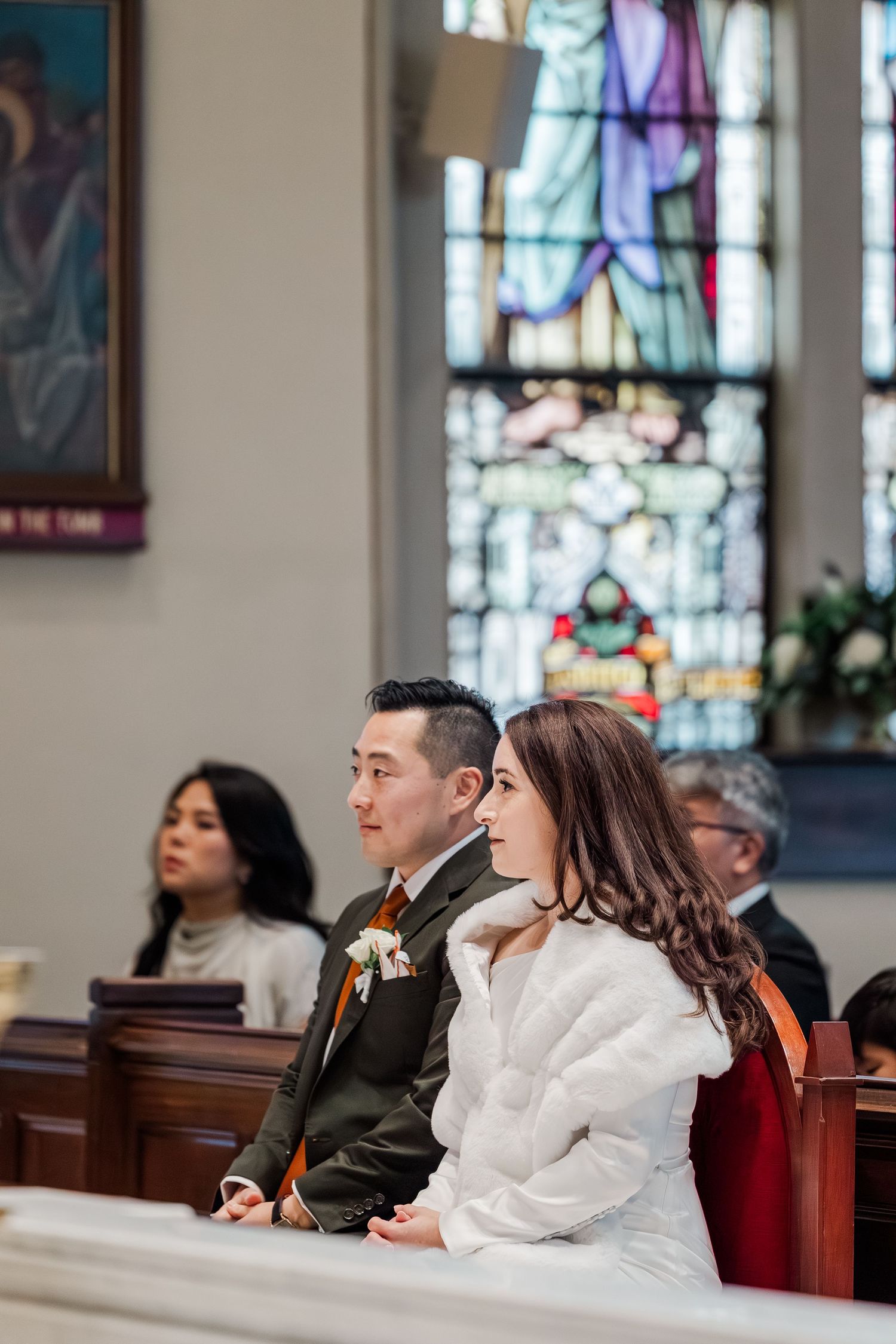 People seated in church pews look up at stained glass windows during a religious service.