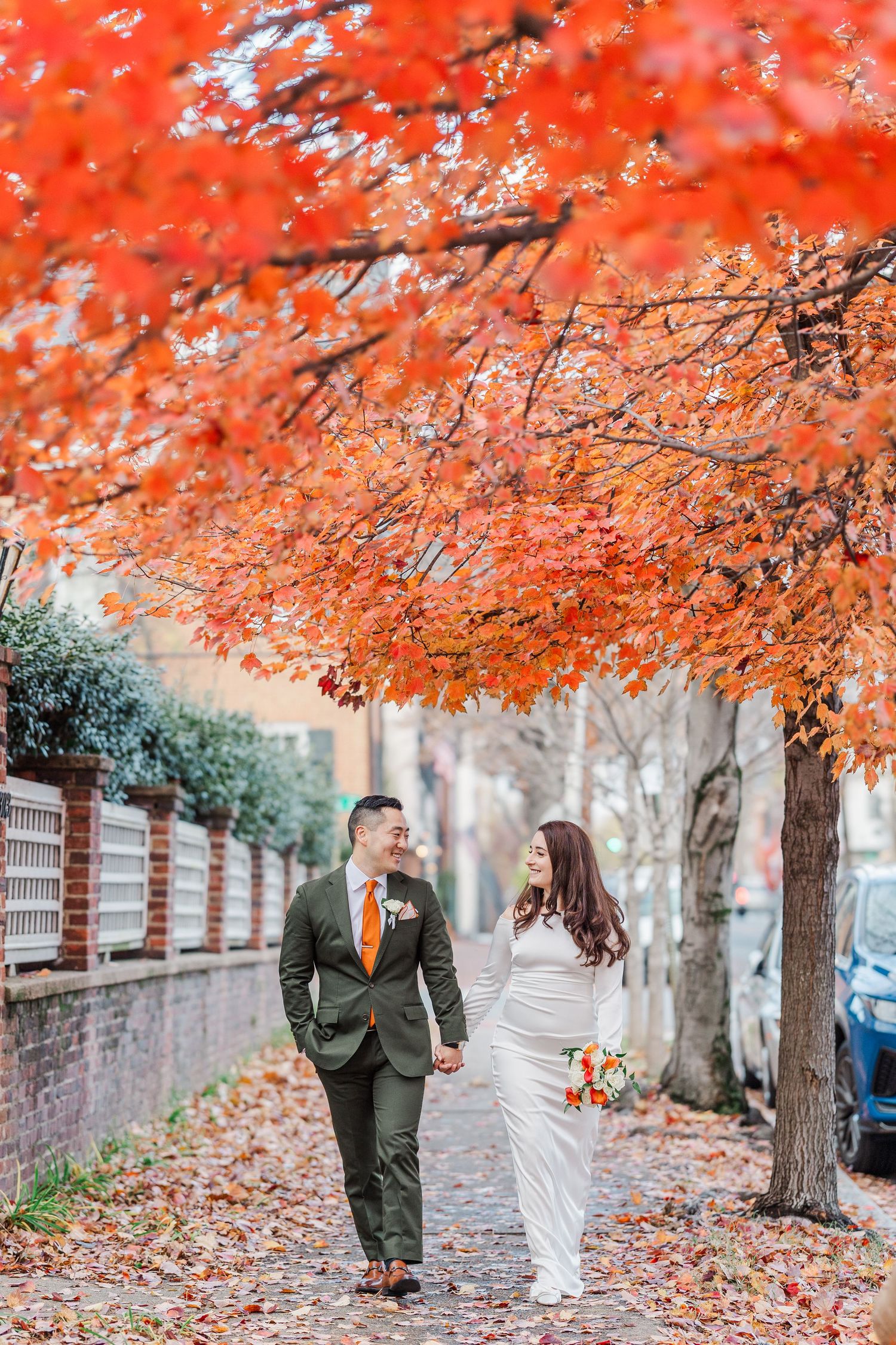 A couple walks down a sidewalk under vibrant orange autumn maple trees with fallen leaves scattered on the ground.