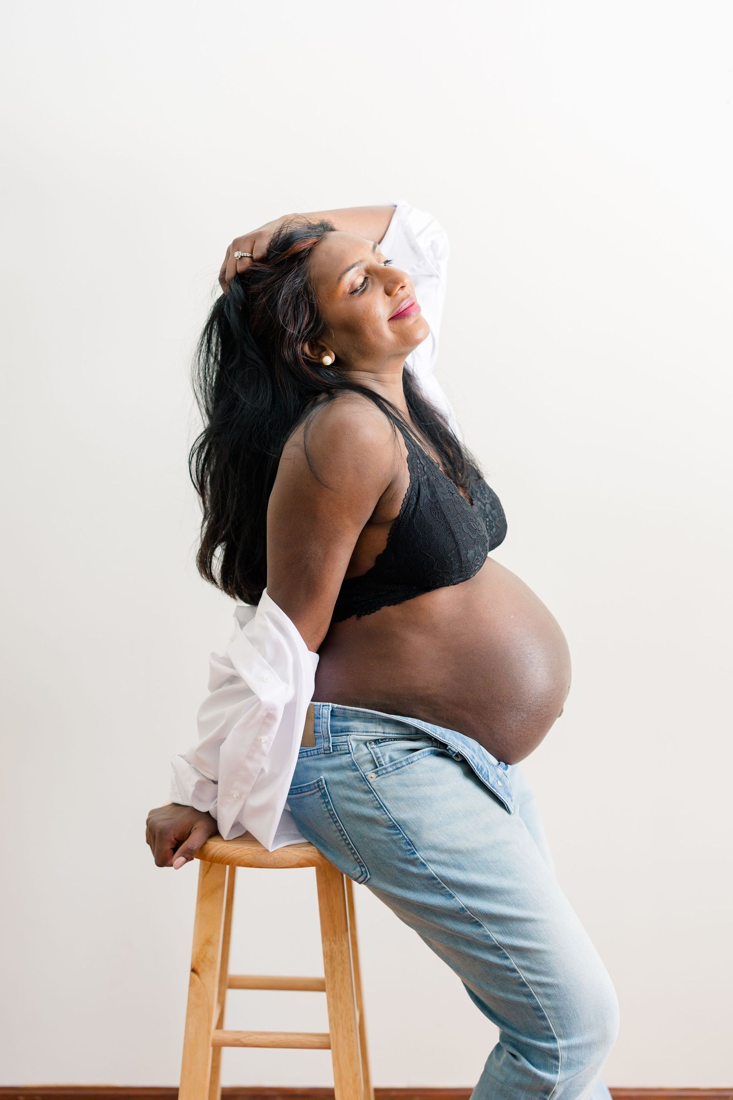 Expectant mother in black top and light jeans leans against wooden stool during maternity photoshoot against white backdrop.
