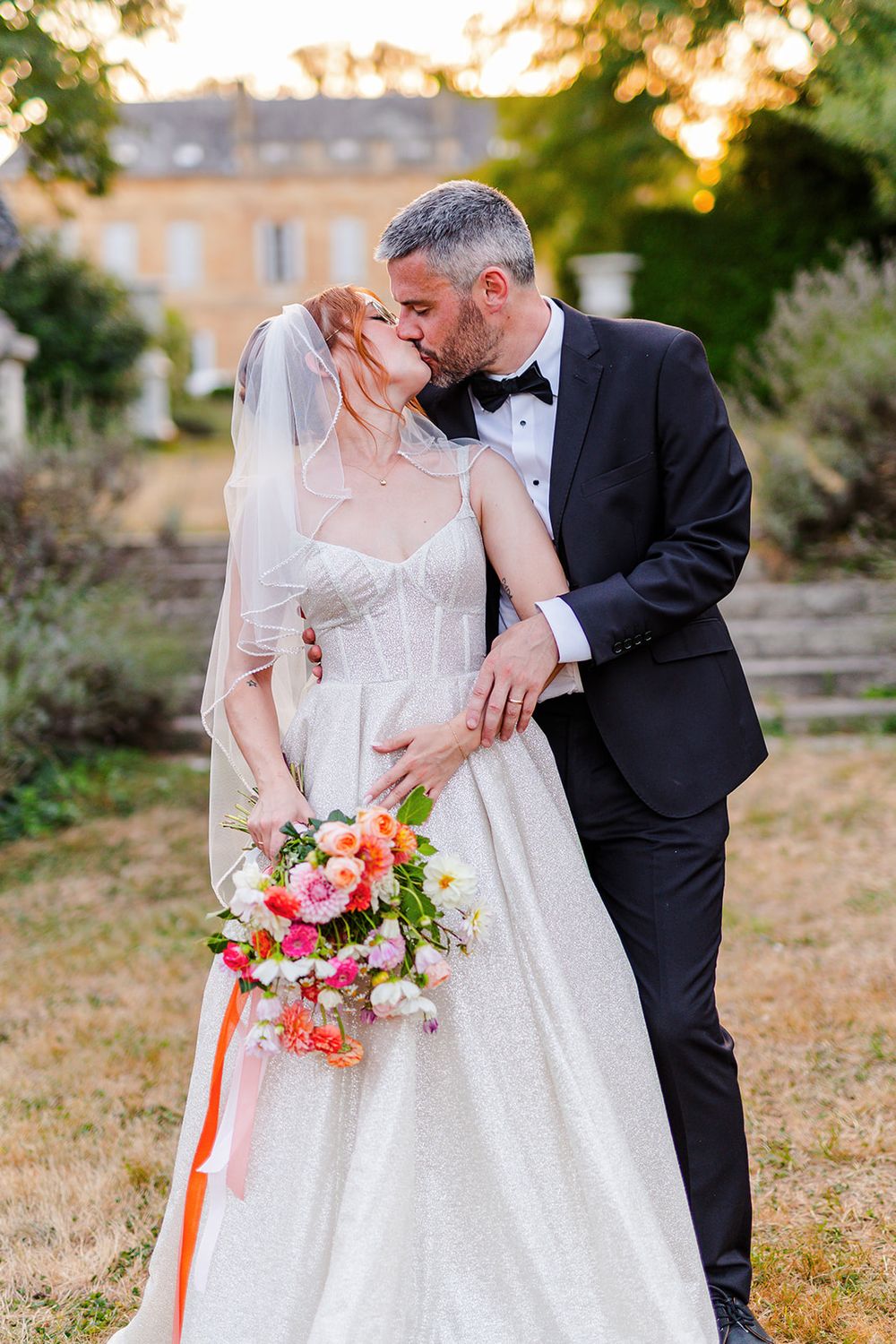 Romantic wedding couple embraces outdoors at sunset holding a vibrant coral and pink floral bouquet.