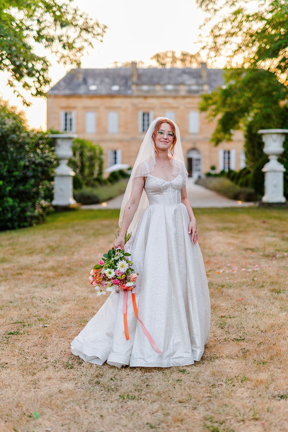 A bride in an elegant white wedding gown holds a vibrant bouquet while standing on a grand estate's grounds at sunset.