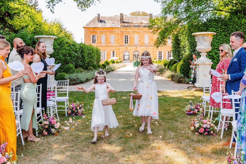 Guests gather on a sunny lawn outside an elegant orange-colored mansion for an outdoor celebration.