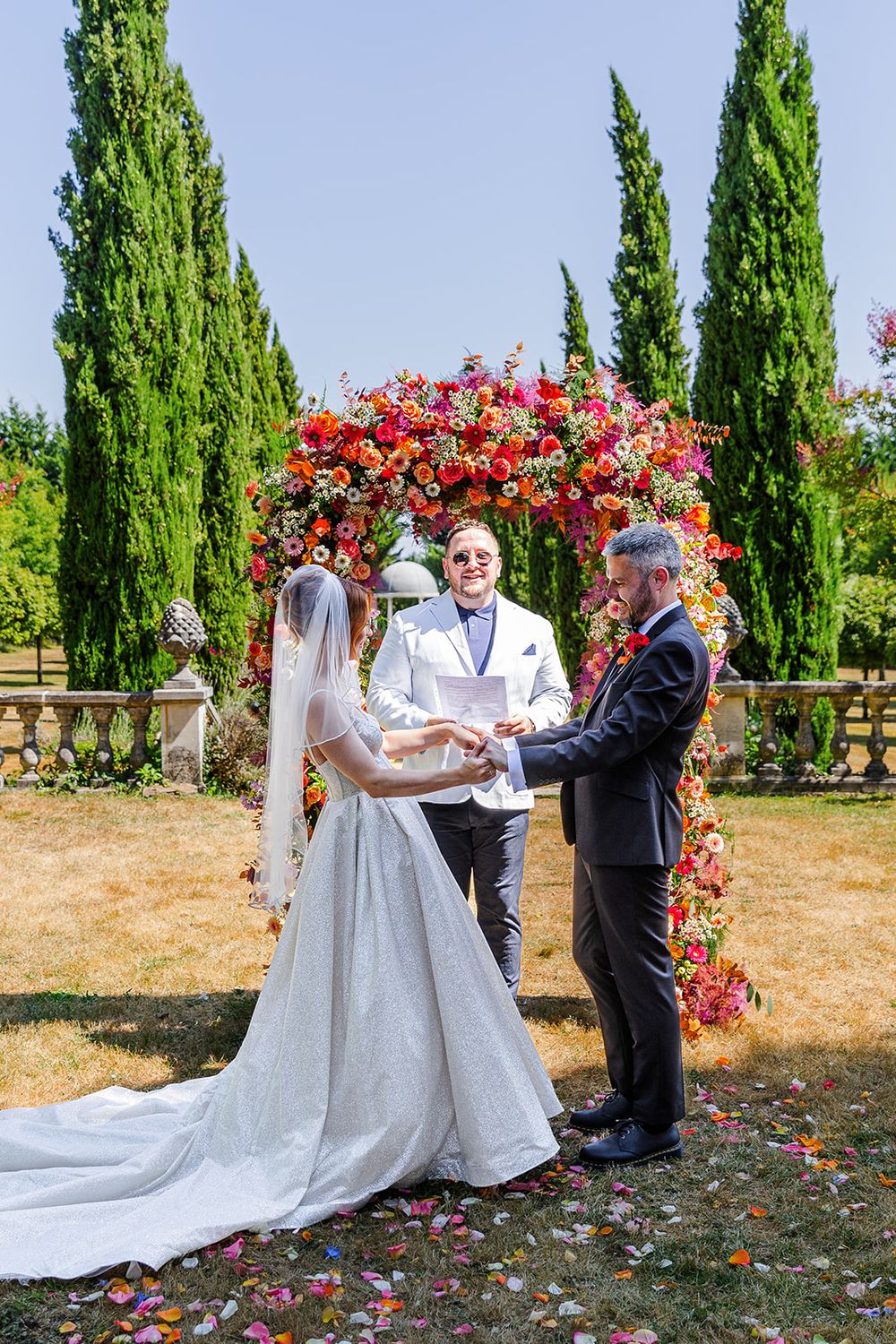 Wedding ceremony taking place under tall cypress trees with red floral decorations.