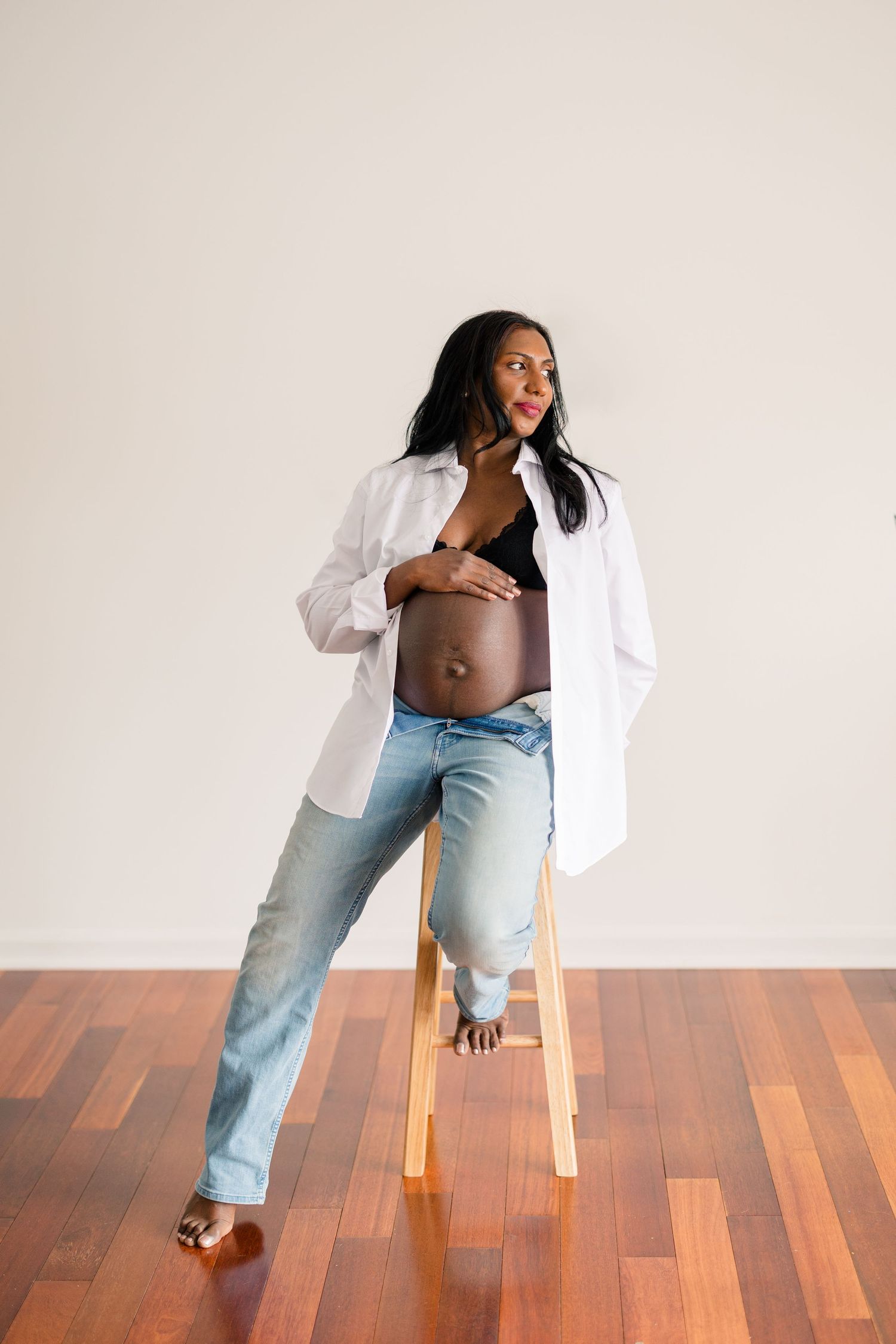 Expectant person sits on wooden stool wearing white button-down shirt and jeans in minimalist maternity photo.