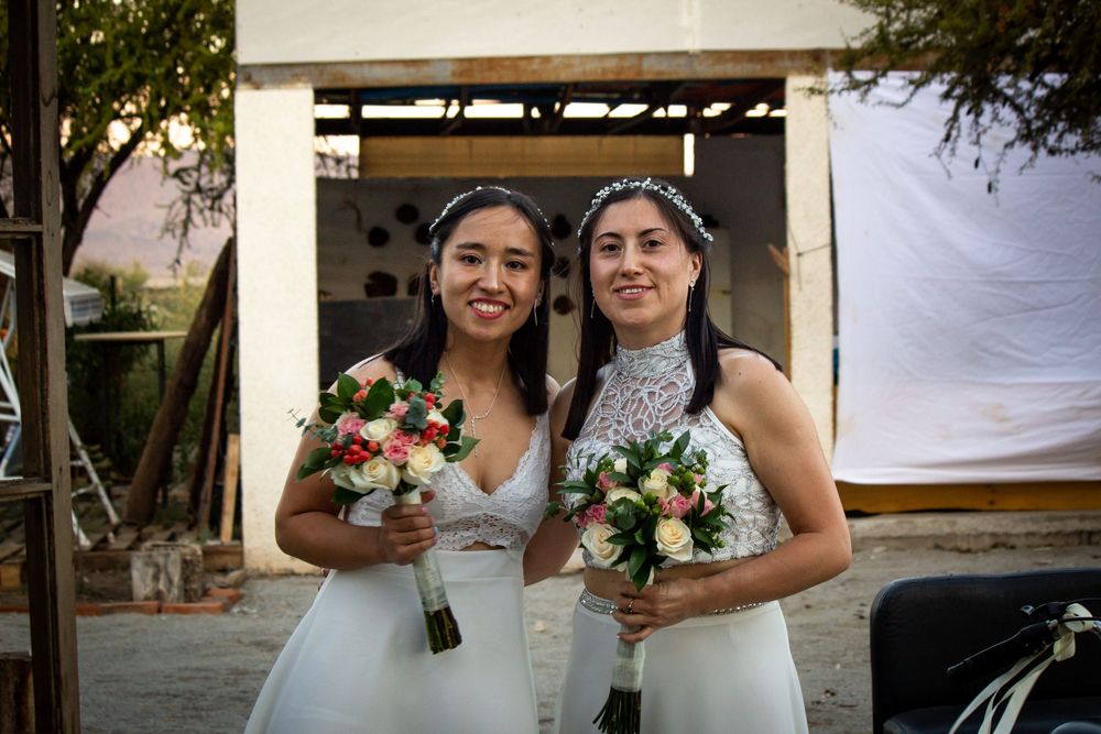 Retrato frontal de pareja de novias con sus ramos de flores, posando sonrientes en el lugar de la recepción al atardecer.