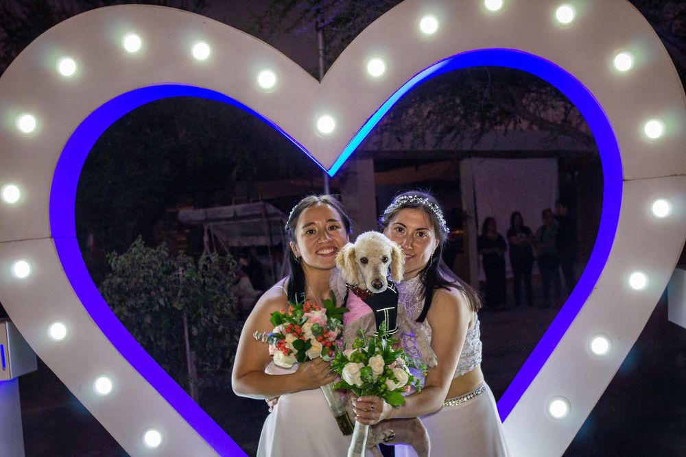 Novias sonrientes posando con su perro dentro de un arco iluminado en forma de corazón; llevan ramos de flores blancas y rosadas.
