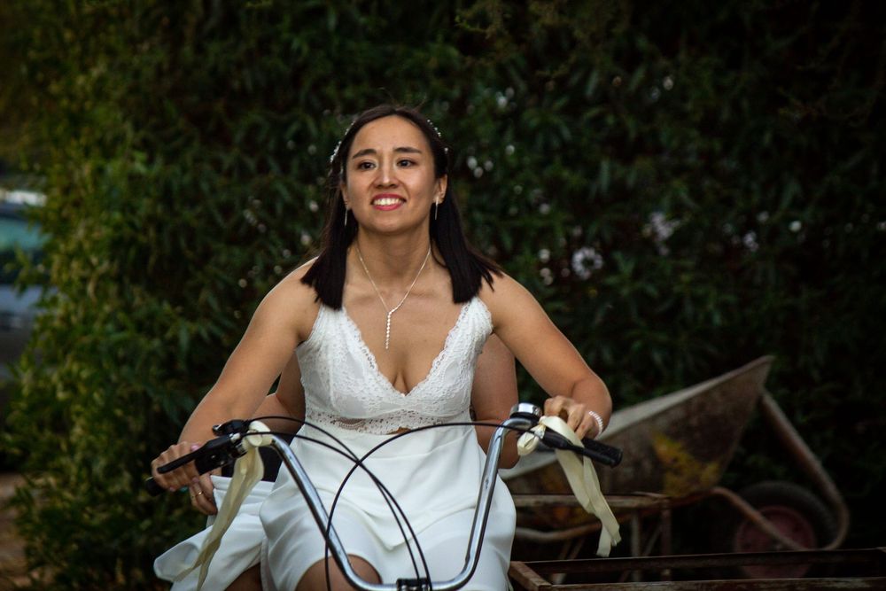 Primer plano de novia sonriente manejando una bicicleta decorada con cintas blancas durante su celebración de boda rústica.