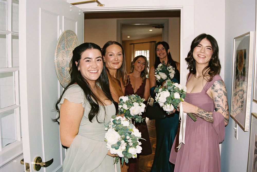 A group of bridesmaids in lavender dresses gather in a hallway holding white and green floral bouquets.