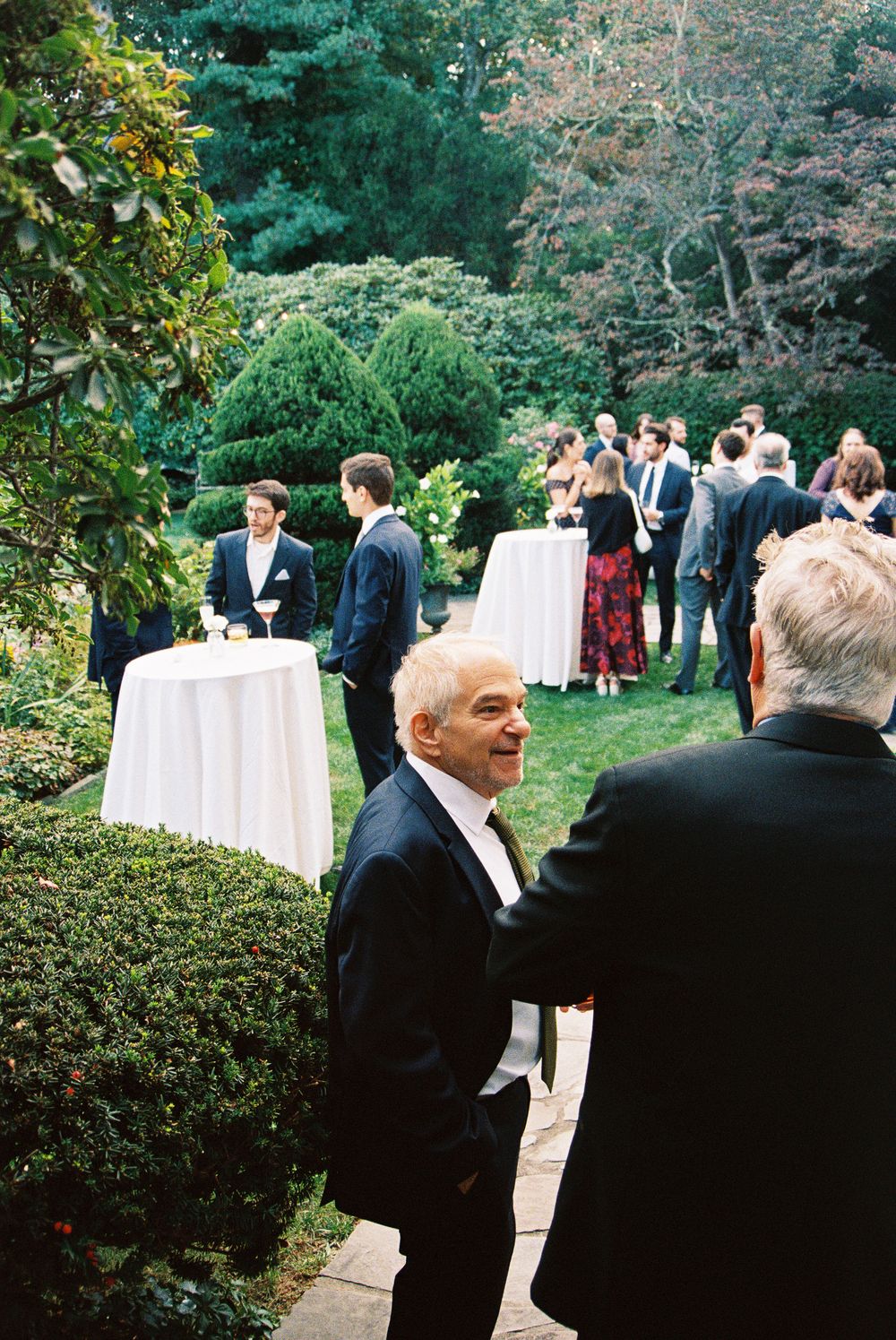Elegant outdoor wedding cocktail hour with guests mingling around white cocktail tables in a lush garden setting.