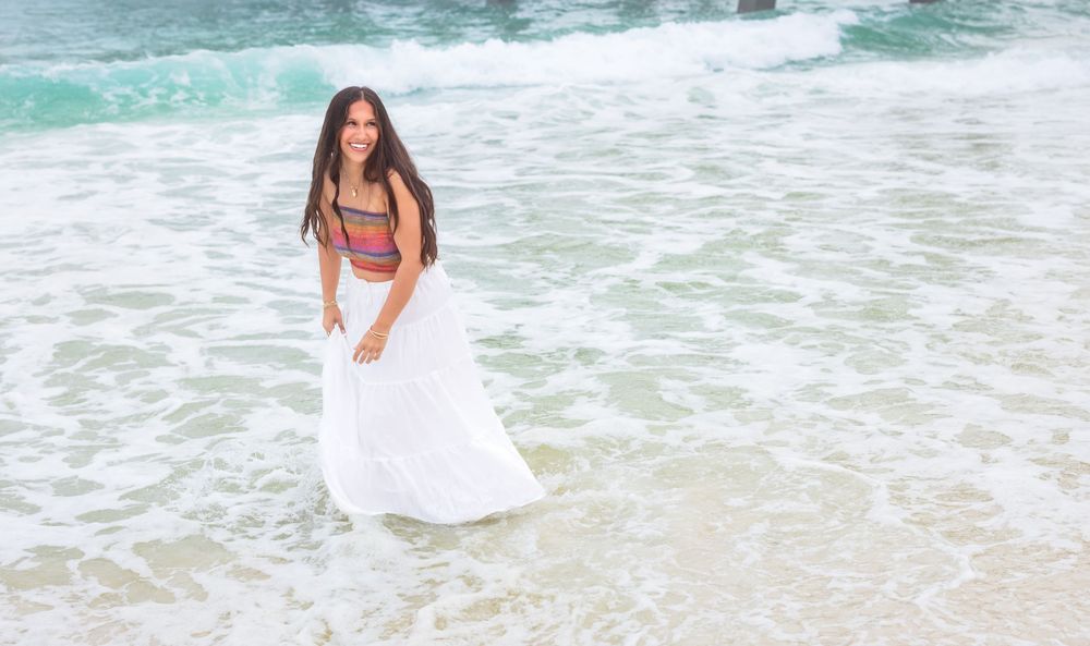 A person wearing a white flowing dress stands in turquoise ocean waves at the beach.