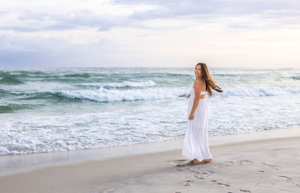A person in a white dress walks along a serene beach with crashing waves and soft pastel sky at sunset.