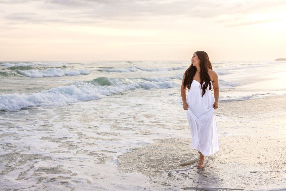 A person in a flowing white dress walks barefoot along the shoreline at sunset as waves roll onto the beach.