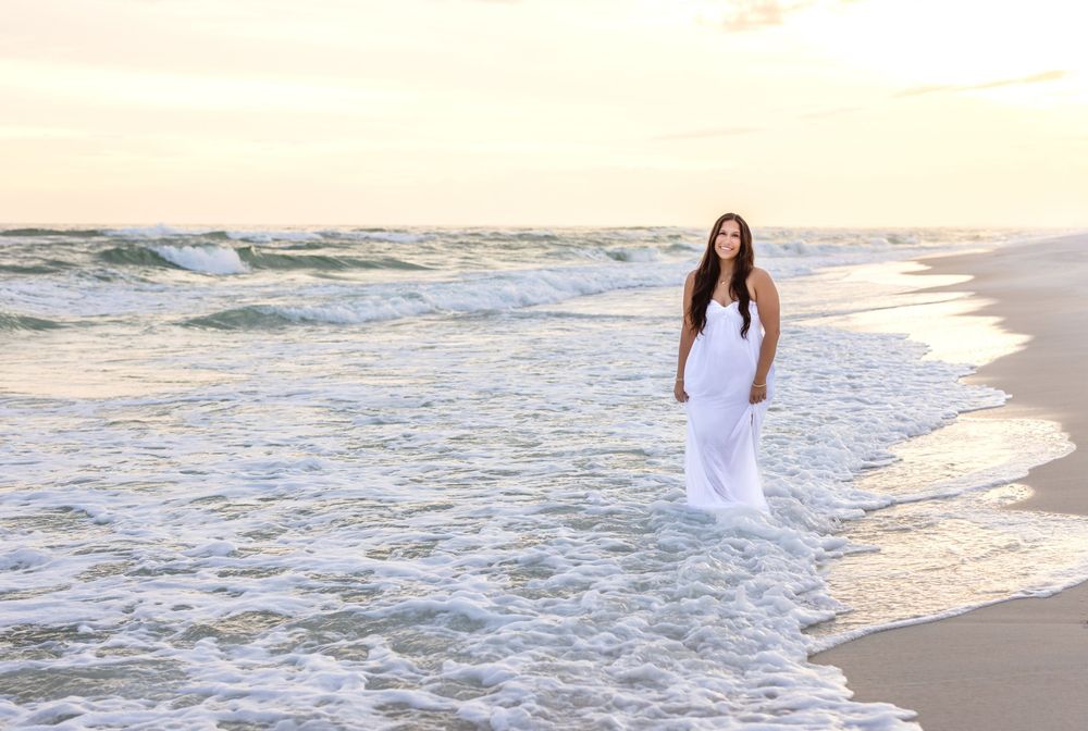 A person in a white dress walks along a sandy beach with ocean waves crashing on the shore at sunset.