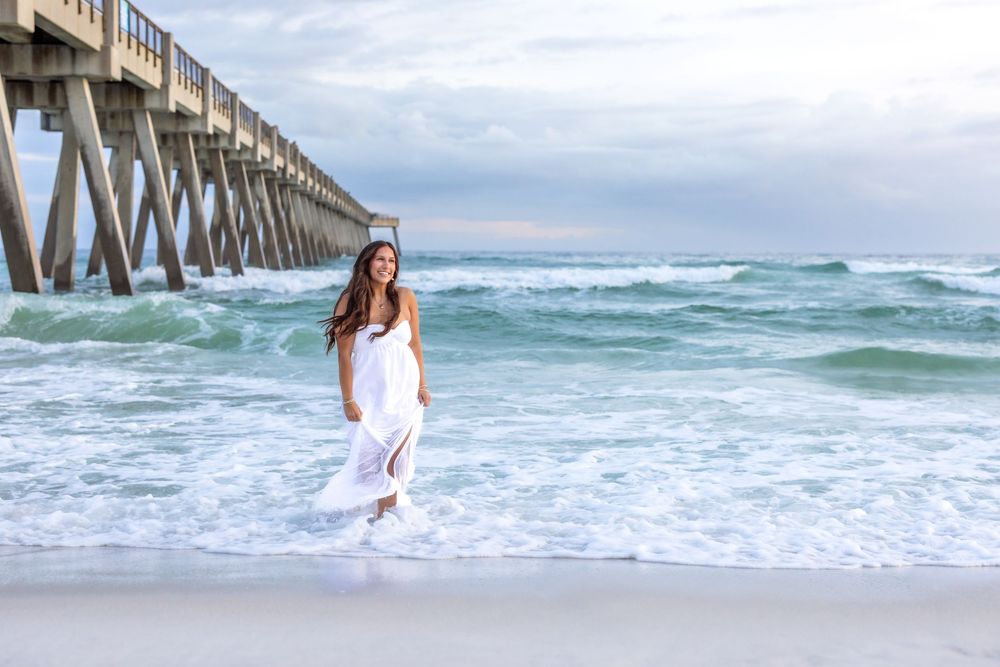 A white-dressed figure walks along the shoreline near a wooden pier as turquoise waves crash against the beach.