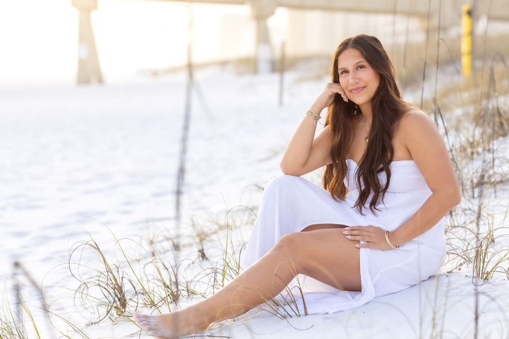 Woman in white dress poses on sandy beach during sunset photo session with dreamy coastal backdrop.