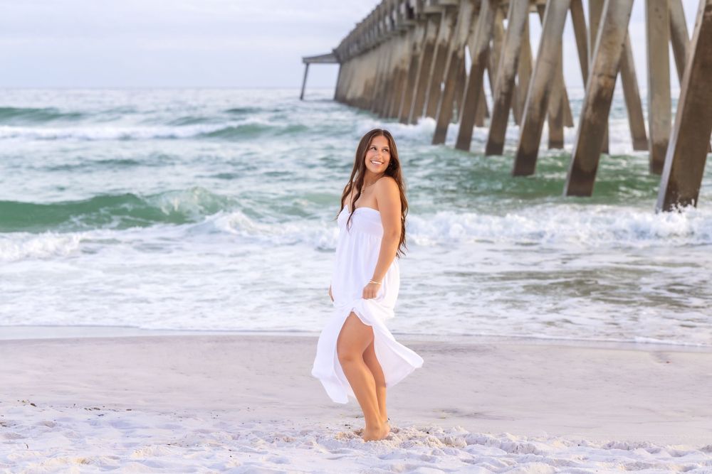 Someone in a flowing white dress poses on the beach near a wooden pier as waves crash in the background.