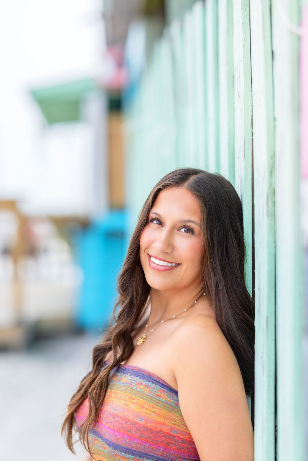 Young woman in colorful strapless top smiles while leaning against mint green wall outdoors.