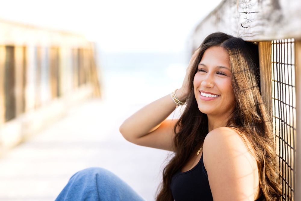A woman in a white dress laughs while sitting on a wooden bench outdoors during a sunny day.