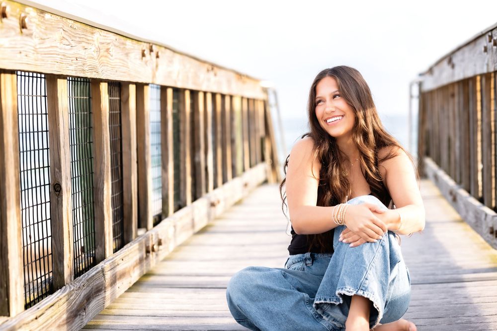 Woman in blue jeans and white top sits casually on wooden boardwalk during golden hour sunset.