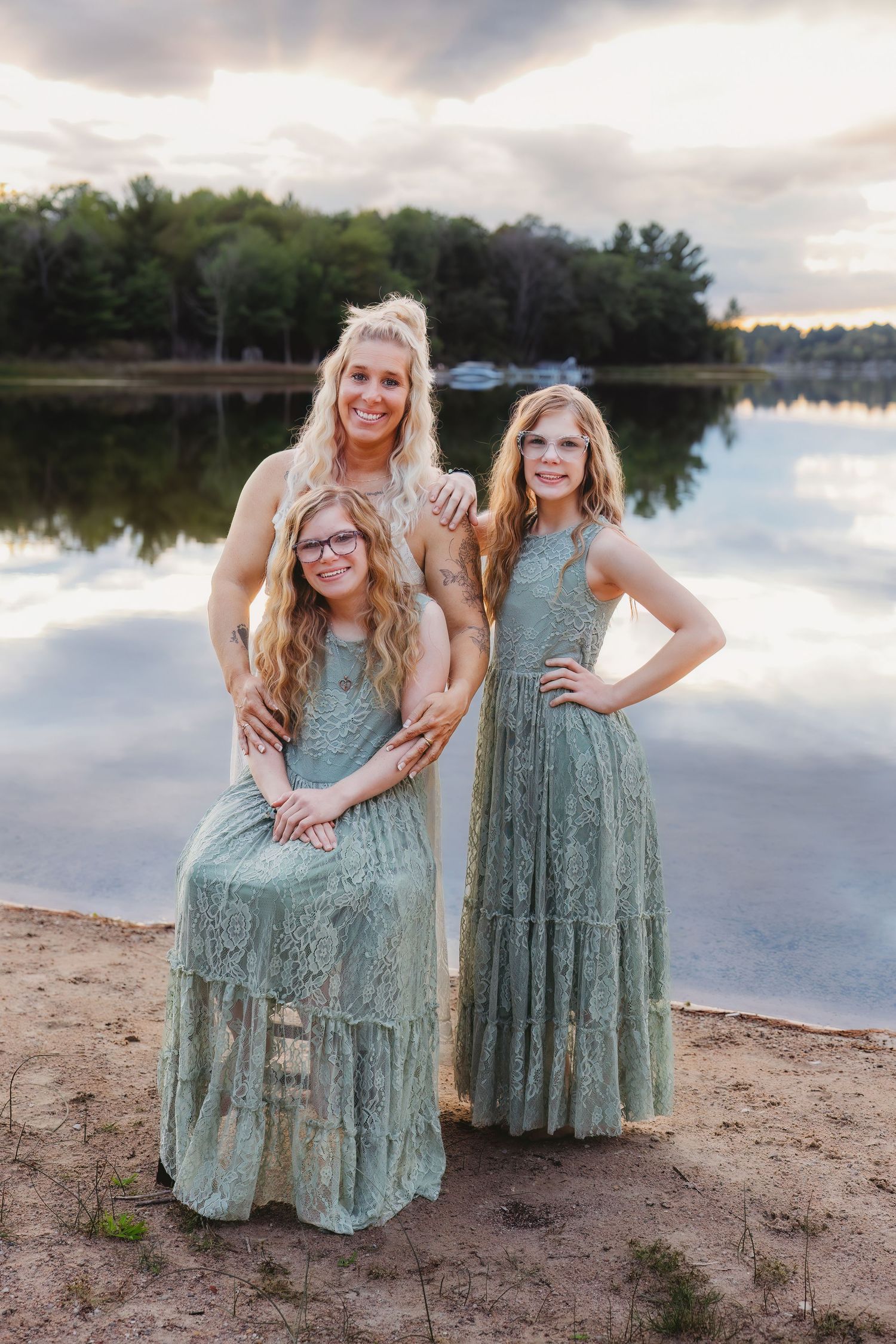 Three people in matching sage green lace dresses pose together on a sandy beach at sunset by a serene lake.
