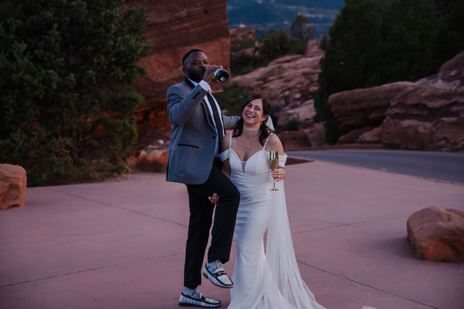 Bride in white gown holds champagne while groom photographs the iconic Red Rocks Amphitheatre at dusk.
