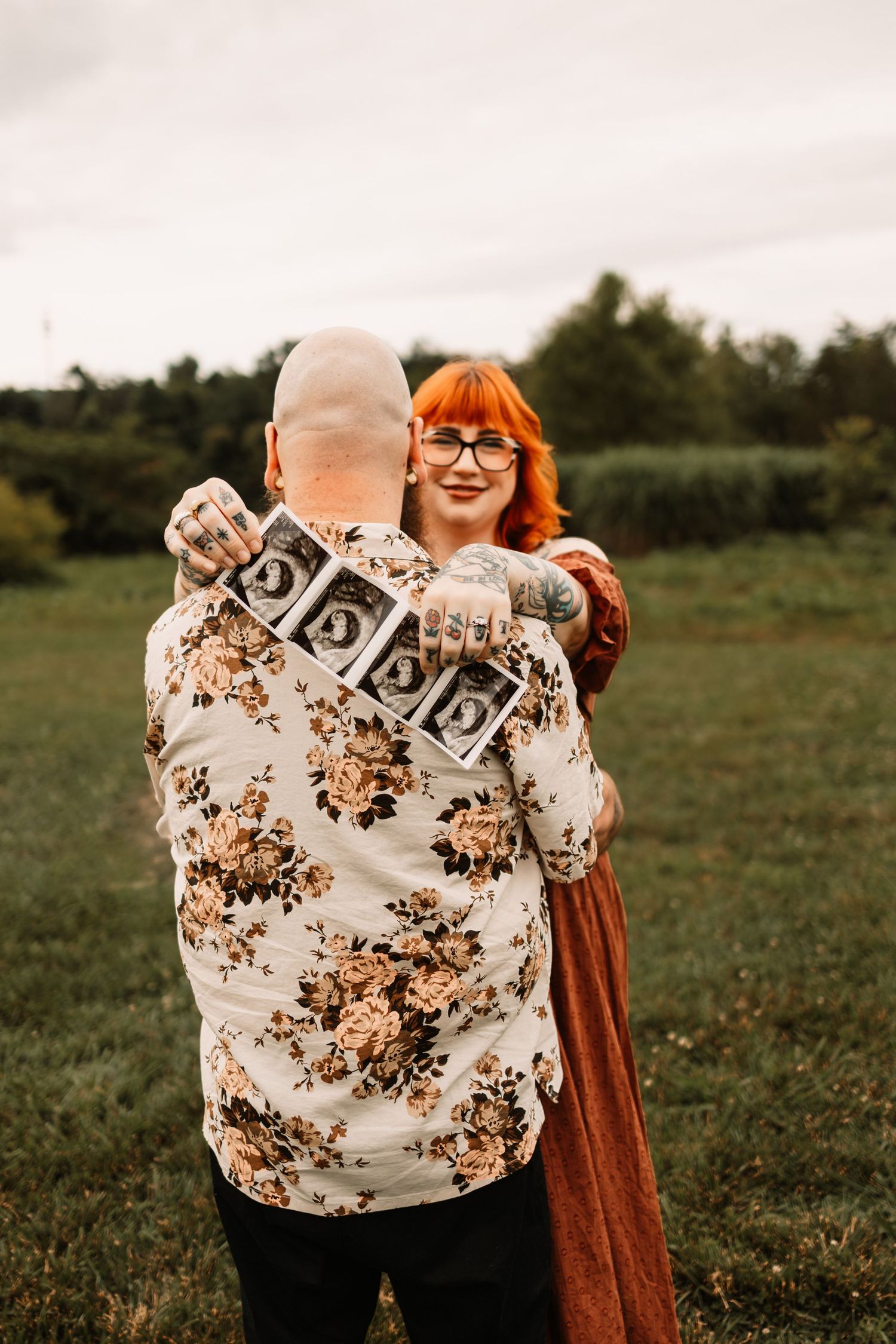 Couple embracing outdoors holding ultrasound photos, woman with red hair and glasses, man in floral shirt with tattoos.