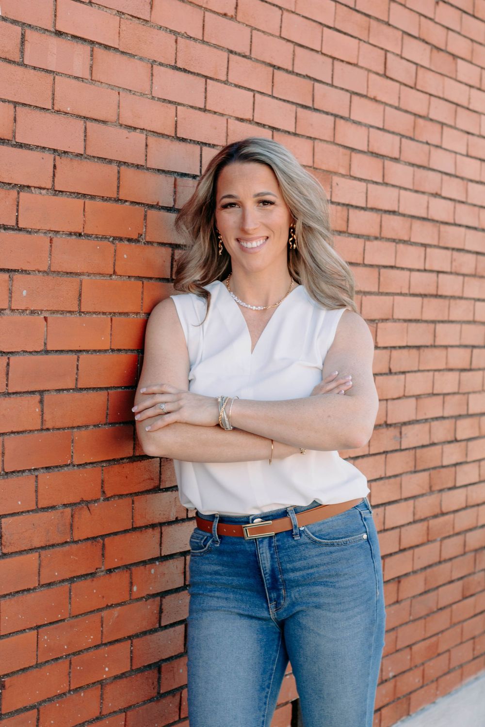 Smiling woman in white sleeveless top and jeans leaning against a red brick wall with arms crossed.