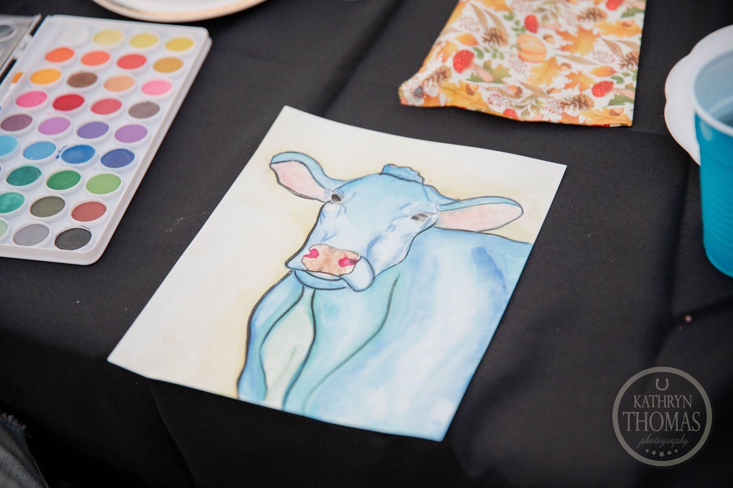Watercolor painting of a blue cow on white paper beside a palette of colorful paints on a black table.