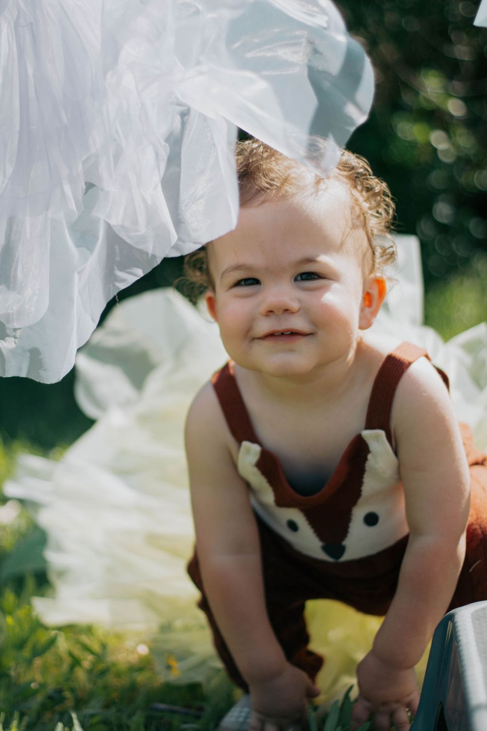 Smiling baby in brown fox romper playing outdoors on green grass with white fabric in sunny garden.