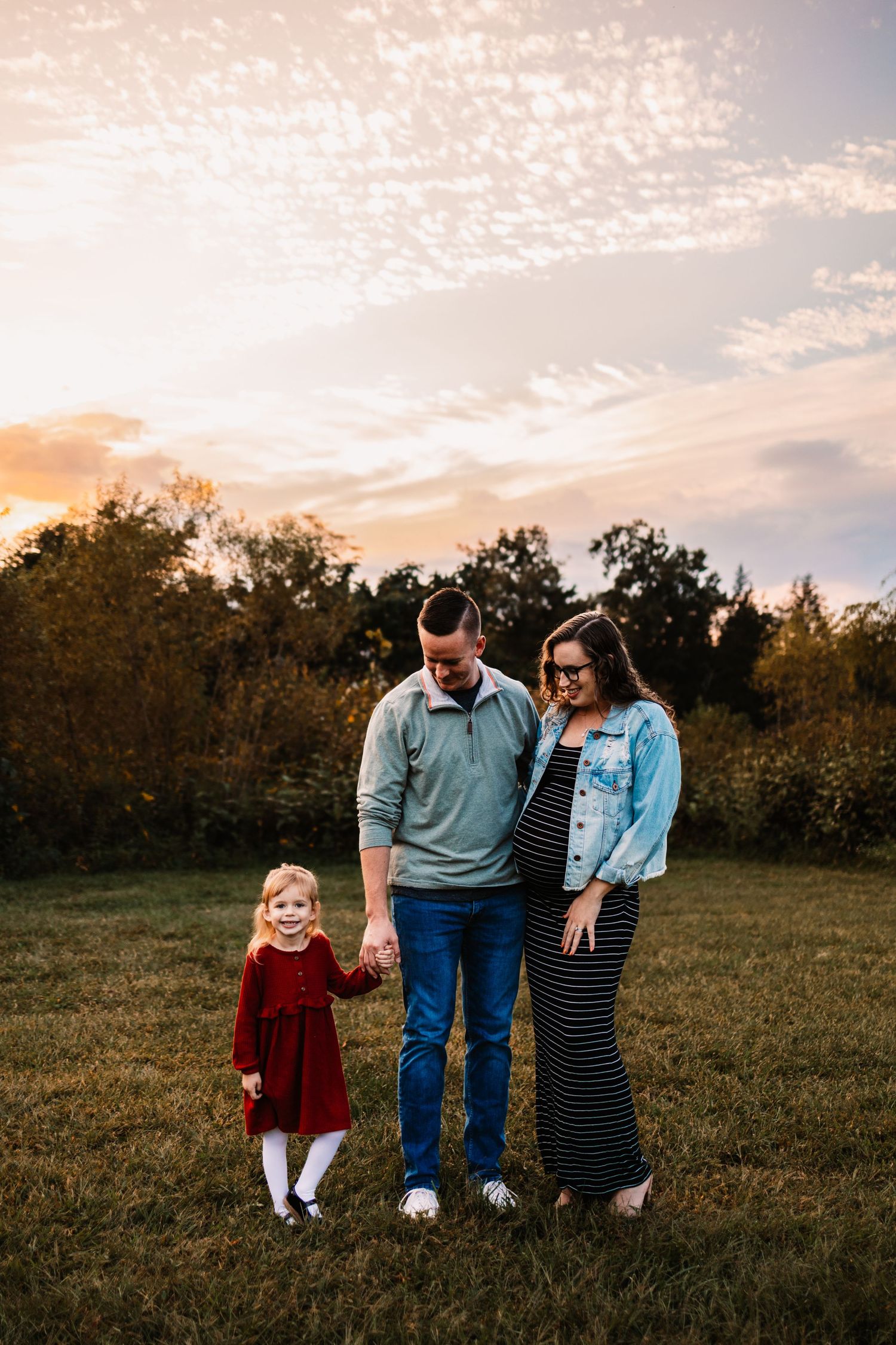 Family of three poses at golden hour in a field, pregnant mom in striped dress, dad in teal sweater, toddler in red dress.