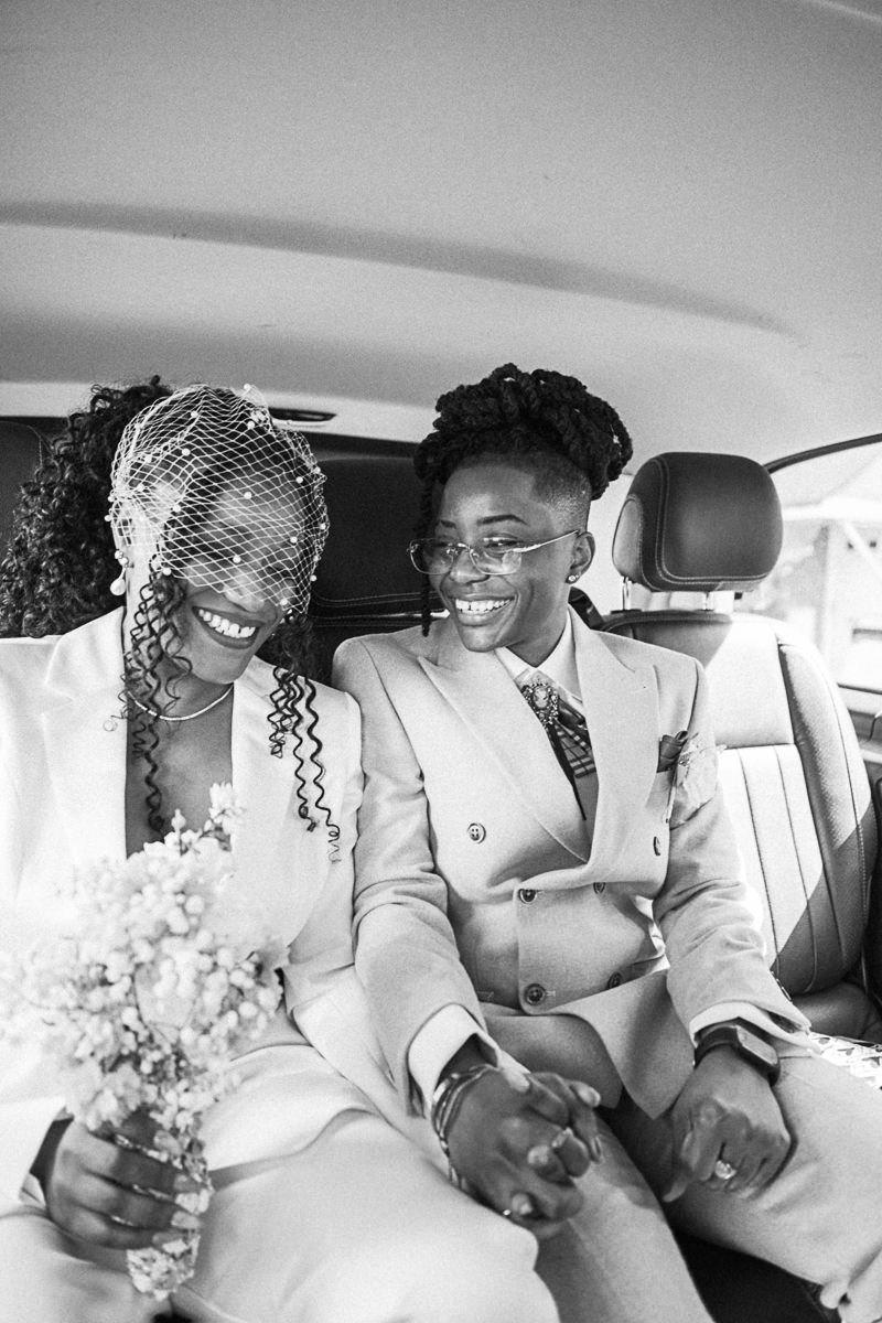 Black and white photo of two brides in suits smiling and holding hands in a car on their wedding day.