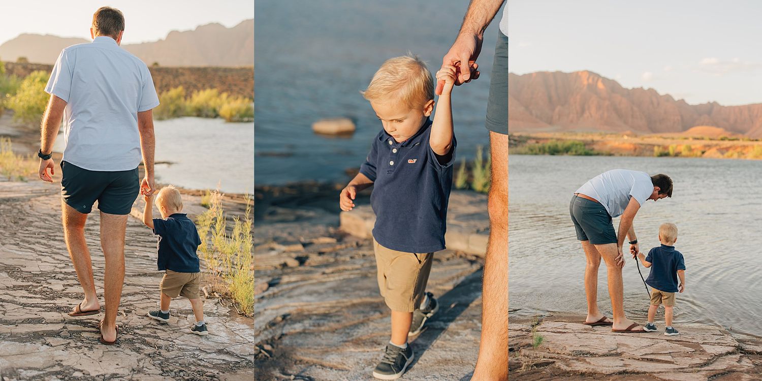 Father and toddler son hold hands walking along rocky riverbank with mountain backdrop at golden hour.